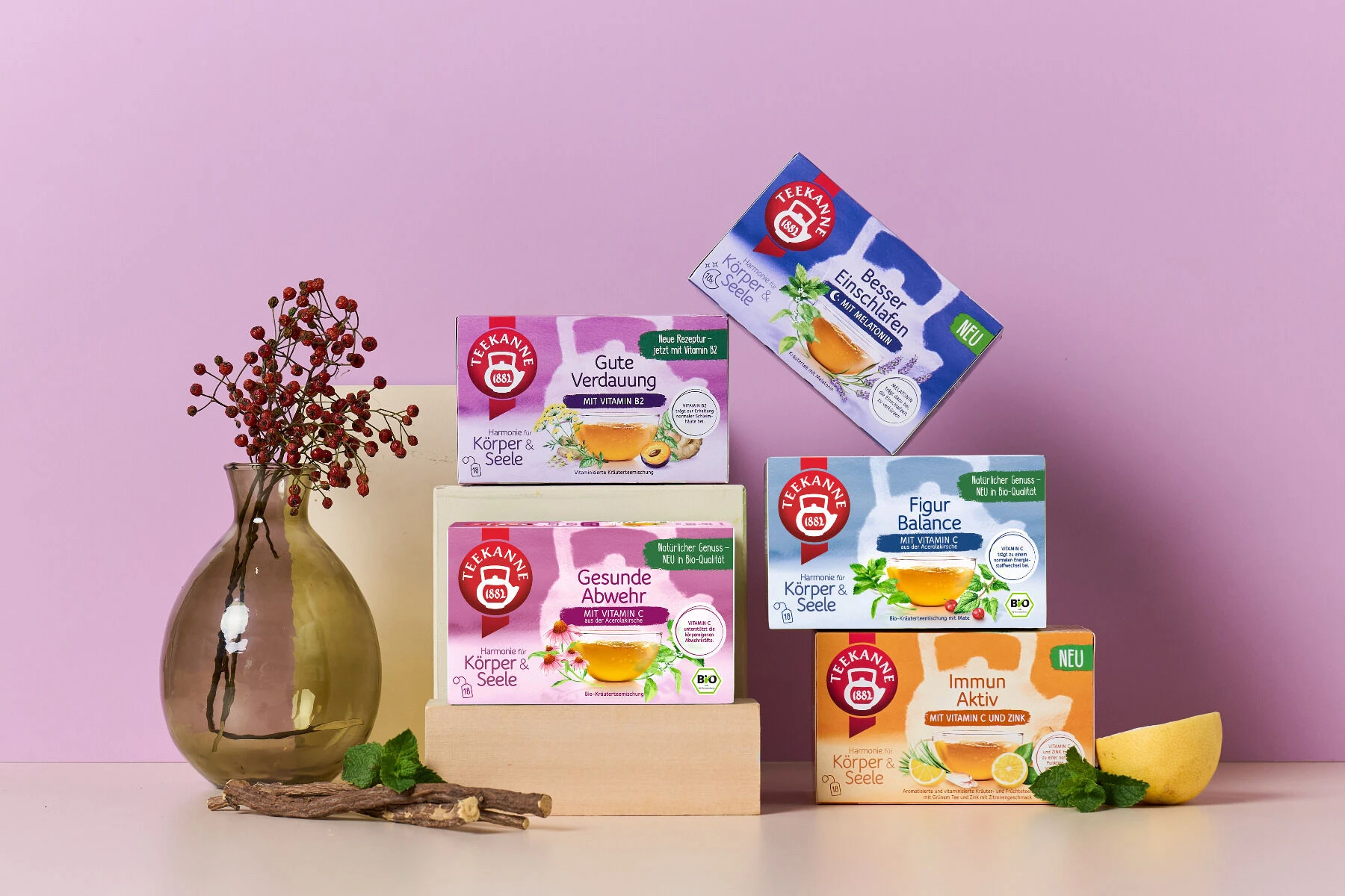 Fünf Schachteln „Harmonie für Körper & Seele Mix & Match Kräutertees“ mit deutschen Etiketten sind auf Holzklötzen vor einem hellvioletten Hintergrund angeordnet, neben einer Glasvase mit roten Beeren, Grünpflanzen und einer Zitronenscheibe in der Ecke.