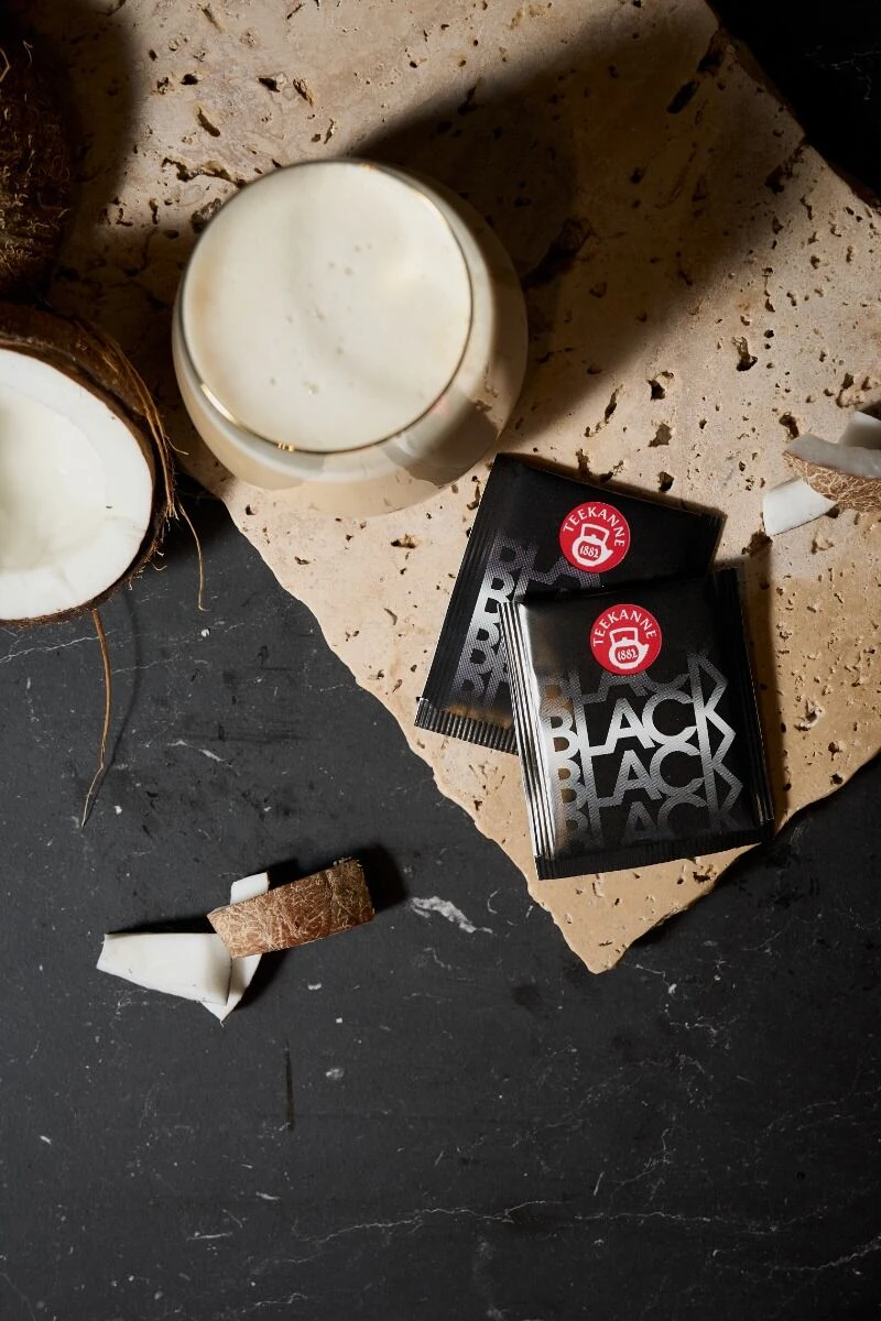 Ein Glas cremiges Getränk „Black Tea Coco & Cream“, zwei Kaffeebeutel „Black Magic“, eine halbierte Kokosnuss und Kokosnussstücke sind auf einer steinernen und dunklen Oberfläche angeordnet.