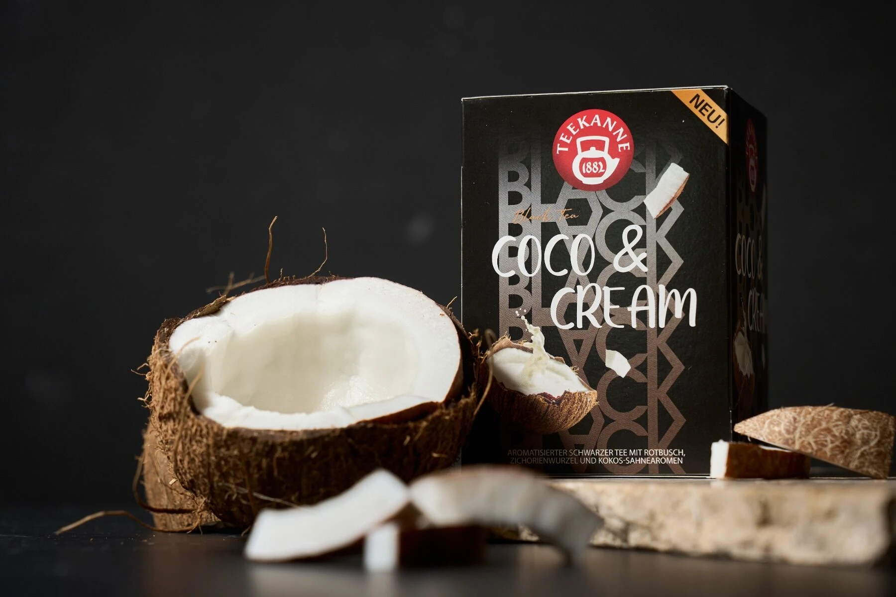 Eine Schachtel Black Tea Coco & Cream steht neben einer halbierten Kokosnuss und Kokosnussstücken auf einer dunklen Fläche mit schwarzem Hintergrund.