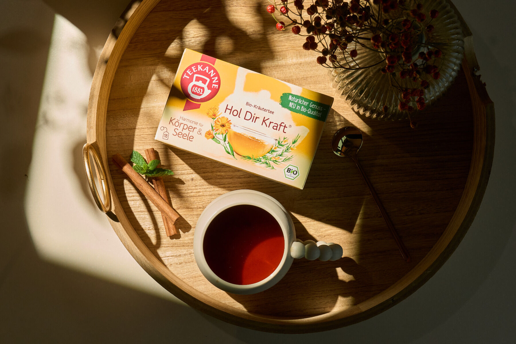 Auf einem runden Holztablett befinden sich eine „Harmonie für Körper & Seele Mix & Match“-Box, eine weiße Tasse Tee, ein goldener Löffel, Zimtstangen und eine Vase mit getrockneten Blumen – wunderschön im Sonnenlicht arrangiert für pure Harmonie.