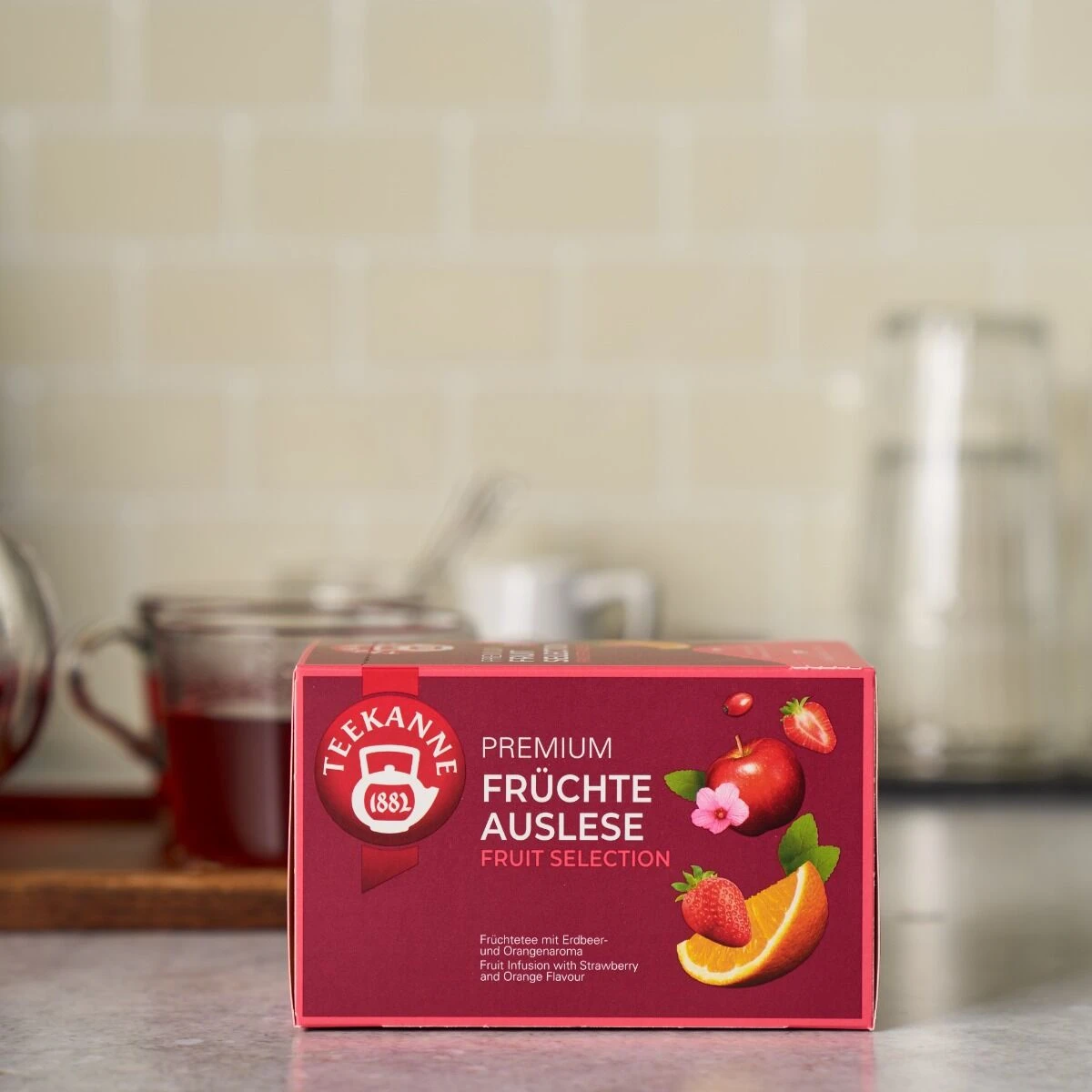 Auf einem Küchentisch steht eine rote Schachtel mit Premium Früchte Auslese, auf der Apfel, Erdbeere und Orange abgebildet sind. In der Nähe stehen eine gläserne Teekanne, eine Tasse Tee und eine Wasserkaraffe.
