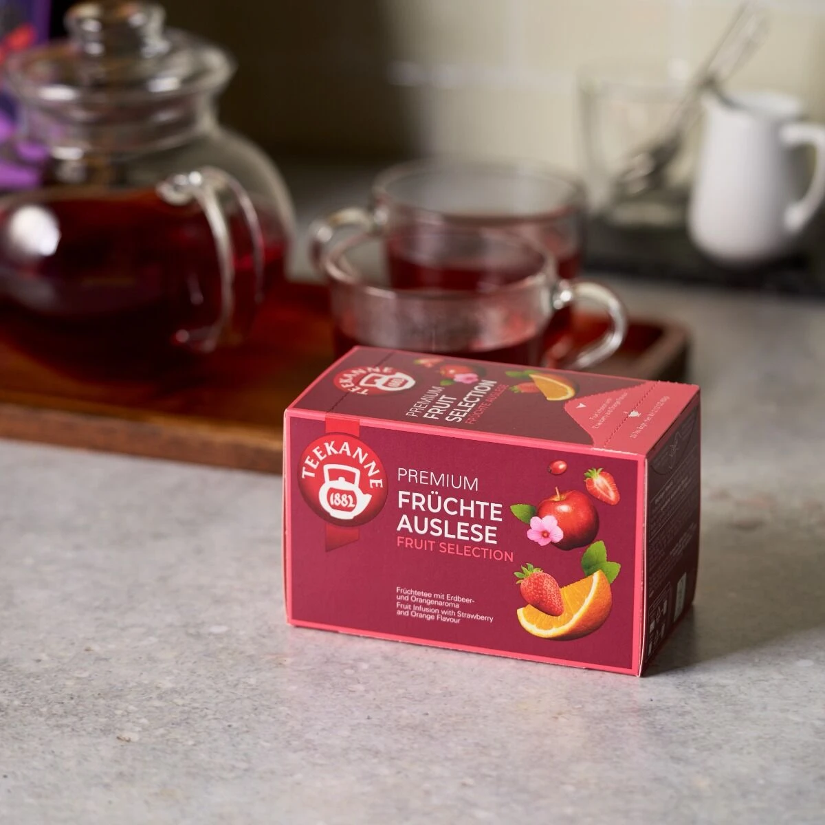 Auf einem Küchentisch steht eine Schachtel Premium Früchte Auslese Tee mit leuchtenden Fruchtbildern, während daneben auf einem Holztablett eine gläserne Teekanne und zwei mit rotem Tee gefüllte Tassen stehen.