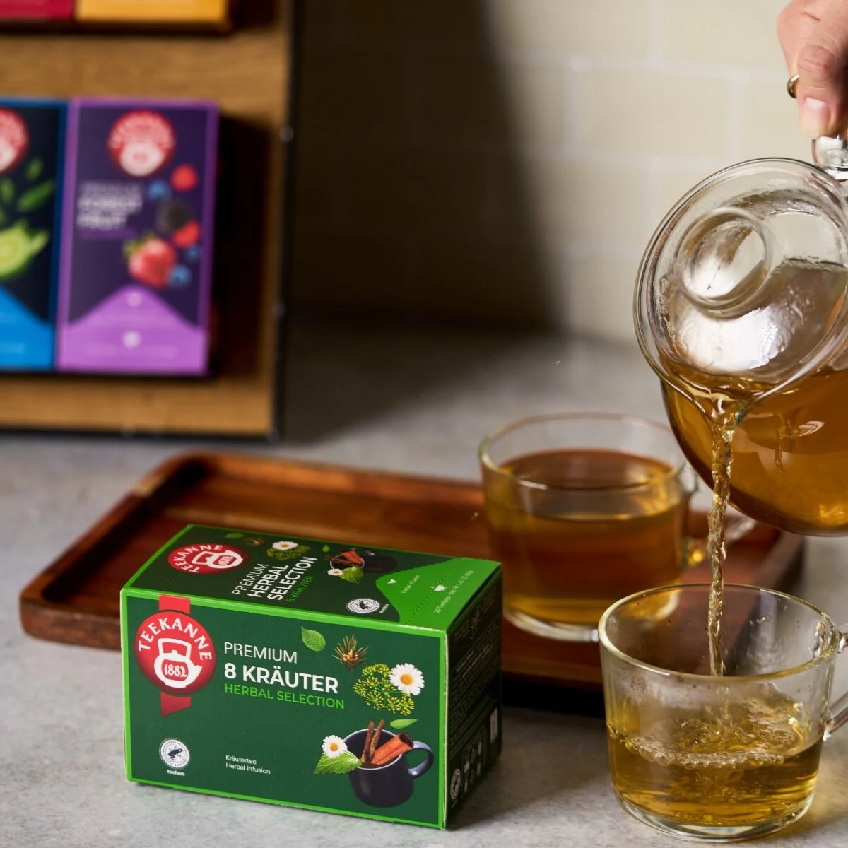 Eine Person gießt Tee aus einer gläsernen Teekanne in eine Tasse. Auf dem Tisch steht eine grüne Schachtel Premium 8 Kräuter, eine Kräuterteemischung mit sorgfältig ausgewählten Kräutern - genießen Sie eine frische Tasse und entdecken Sie sie jetzt!.