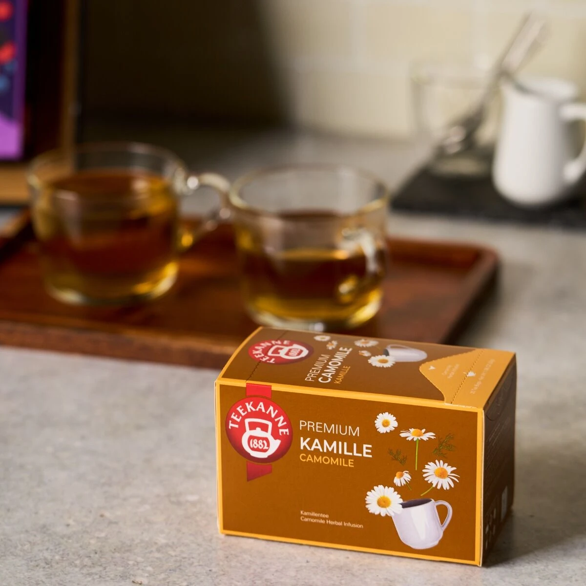 Eine Schachtel Premium Kamille steht auf einem Küchentisch, begleitet von zwei Glastassen mit Kamillentee und einem Holztablett im unscharfen Hintergrund.