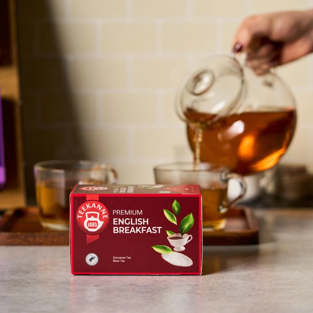 Auf dem Tresen steht eine Schachtel Premium English Breakfast Tee, der aus einer gläsernen Teekanne in eine durchsichtige Tasse gegossen wird, während im Hintergrund zwei einladende Tassen Premium English Breakfast auf einem Holztablett stehen.