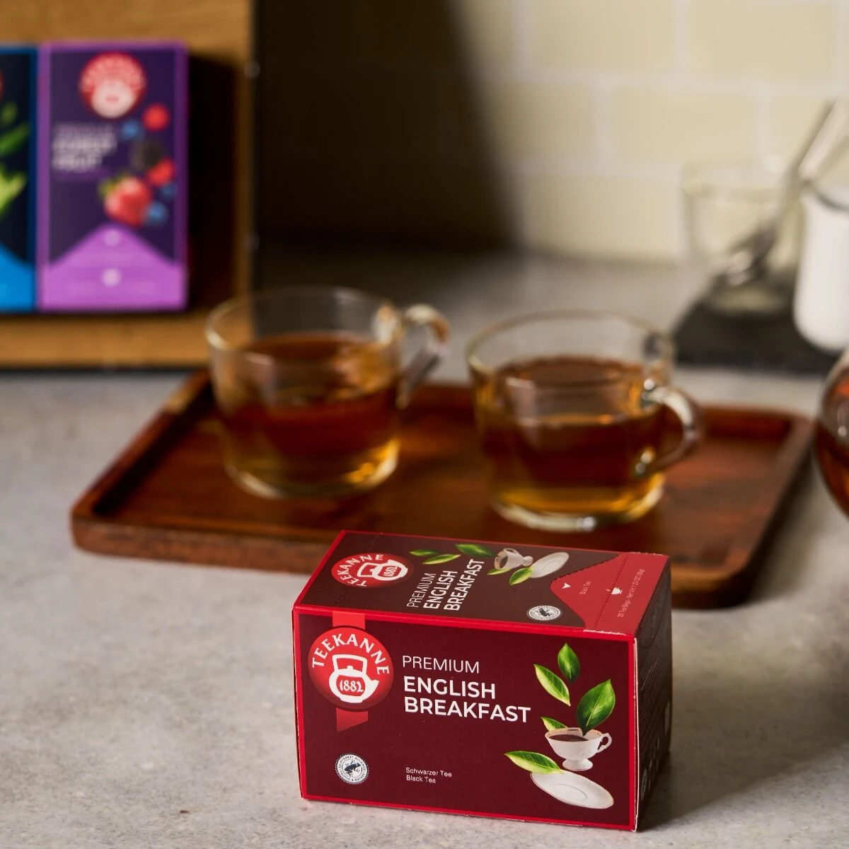 Eine Schachtel Premium English Breakfast Tea steht auf einem Küchentisch neben zwei Glastassen mit Tee auf einem Holztablett, während andere Teeschachteln im Hintergrund verschwimmen.