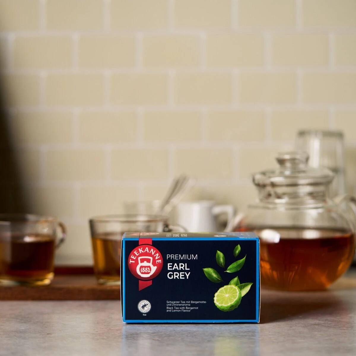 Eine Schachtel Premium Earl Grey steht auf einem Küchentisch, im Hintergrund zwei Glastassen und eine Teekanne mit Tee. Auf der Verpackung sind eine Limette und Teeblätter abgebildet.