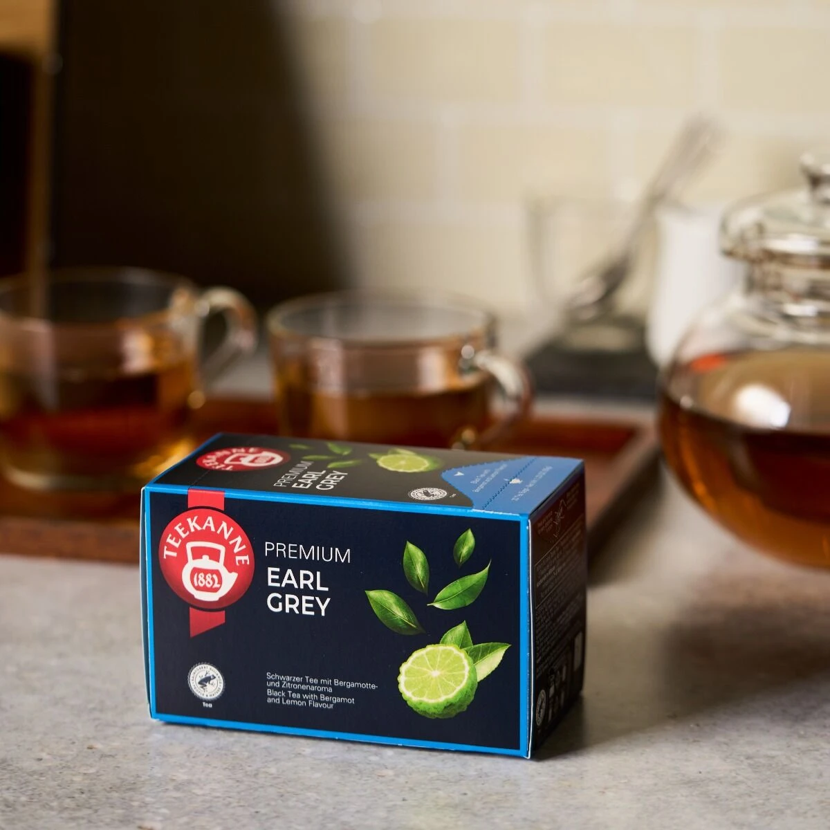 Auf der Arbeitsplatte steht eine Schachtel Premium Earl Grey Tee, im Hintergrund stehen zwei Glastassen und eine mit aromatischem Tee gefüllte Teekanne.