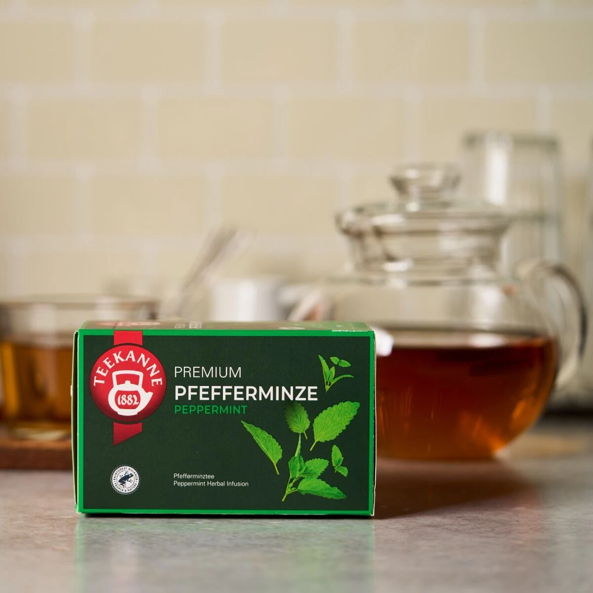 Eine Schachtel Premium Pfefferminze-Tee steht auf einem Küchentisch. Im unscharfen Hintergrund sind eine gläserne Teekanne und eine Teetasse zu sehen. Auf der Vorderseite der grünen Schachtel sind Minzblätter abgebildet.