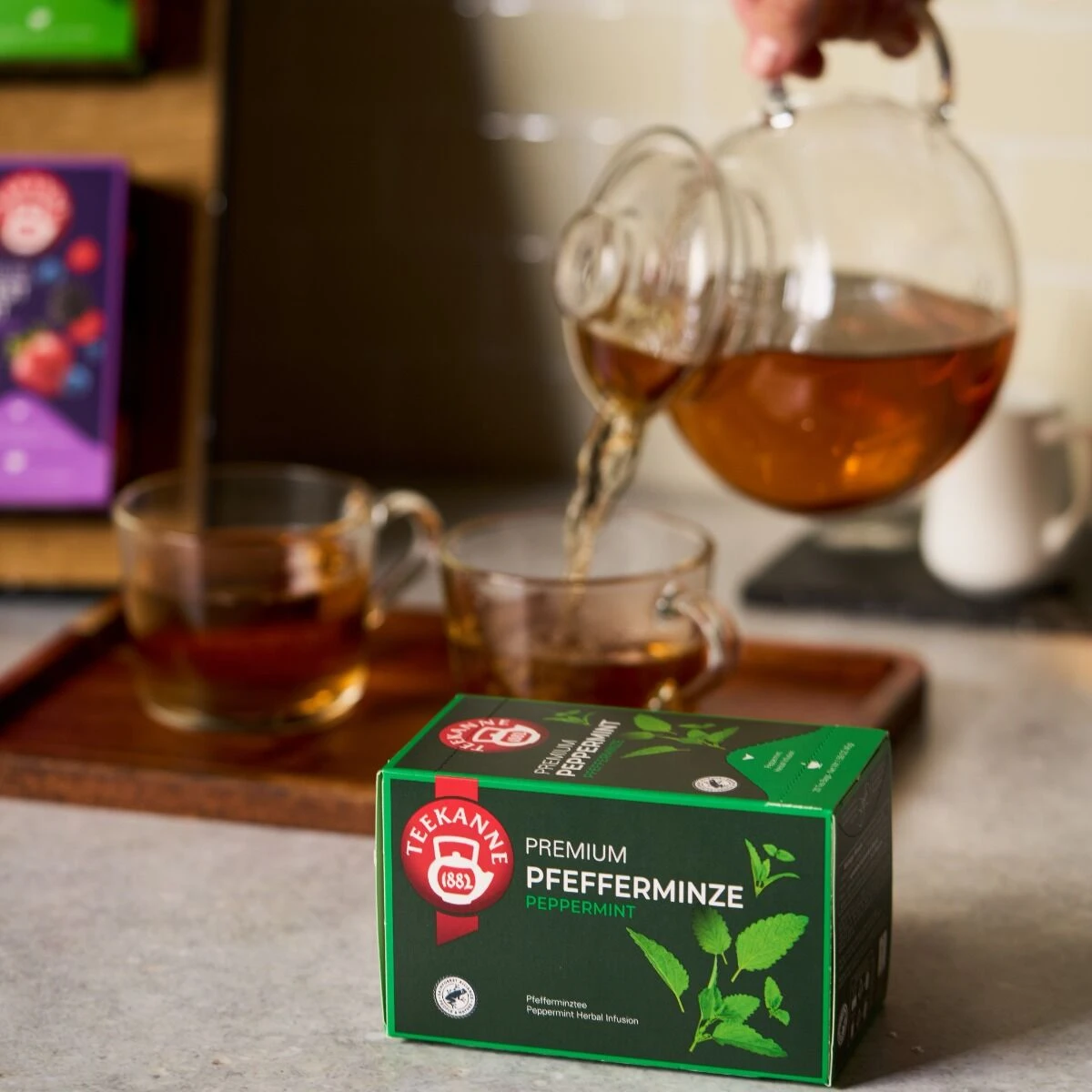 Eine Schachtel Premium Pfefferminze-Tee steht auf dem Tresen, während im Hintergrund der Tee aus einer gläsernen Teekanne in eine klare Tasse auf einem Holztablett gegossen wird.