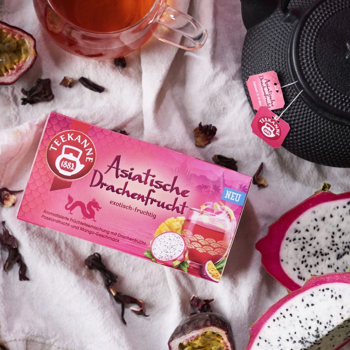 Eine Schachtel Asiatische Drachenfrucht-Tee steht auf einem Tisch, umgeben von geschnittenen Drachenfrüchten, saftigen Passionsfrüchten, einer schwarzen Teekanne, einer Tasse Tee und verstreuten Hibiskusblättern.