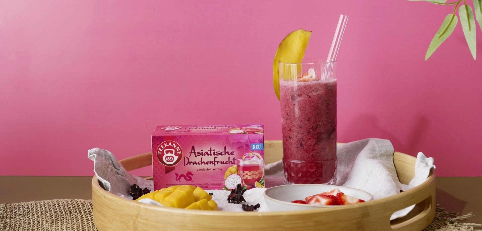 Auf einem Holztablett stehen ein Glas Asiatische Drachenfrucht-Smoothie mit Strohhalm und Mangoscheibe, eine Schachtel Tee, eine Schale mit Fruchtstücken sowie frische Mango und Drachenfrucht, alles vor einem leuchtend rosa Hintergrund.