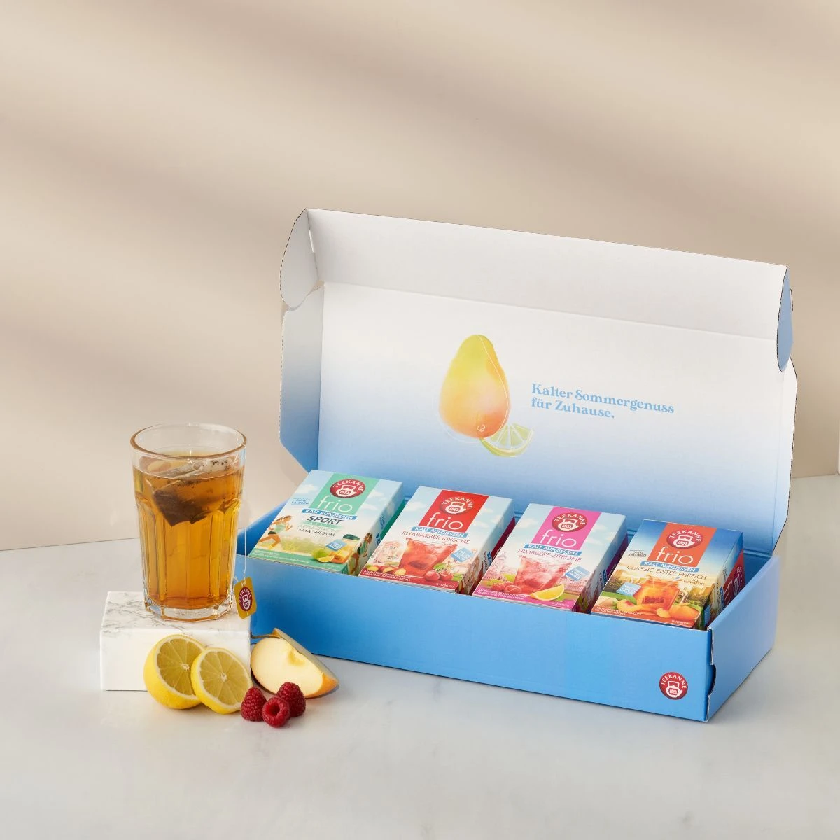 Eine blaue Frio Geschenkbox mit vier Teeaufgüssen steht geöffnet neben einem Glas Frio Eistee mit Eiswürfeln und einer Zitronenscheibe. Zitronen- und Pfirsichscheiben sind auf einer weißen Fläche neben der Box angeordnet.