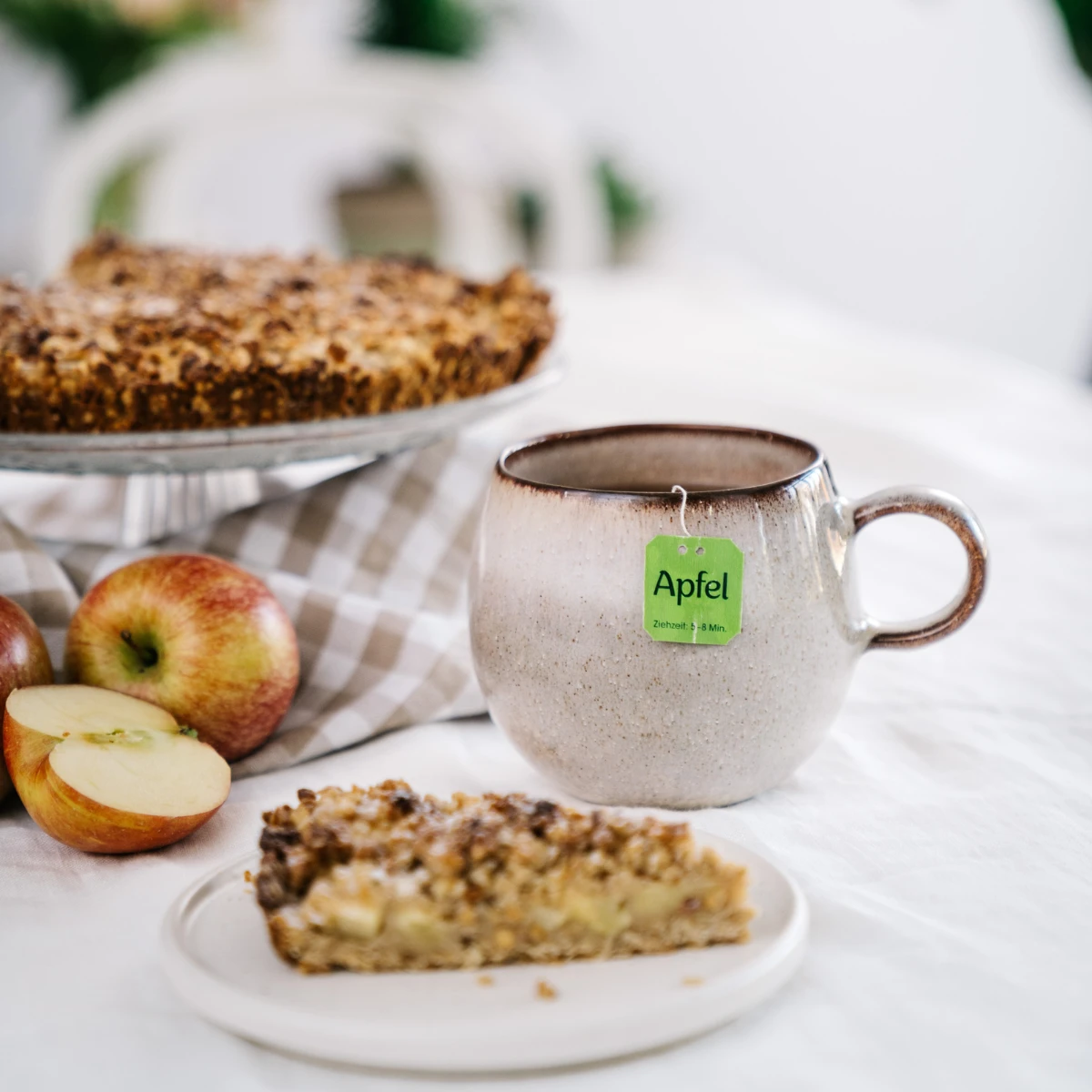 Eine Keramiktasse mit Apfeltee steht neben einem Stück Apfelstreuselkuchen auf einem weißen Teller, ganzen Äpfeln und einem größeren Kuchen im Hintergrund auf einem karierten Tuch.
