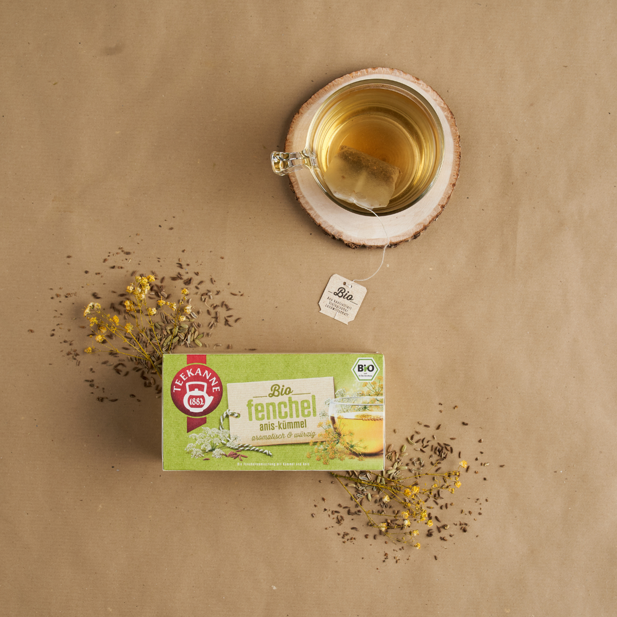 Eine Tasse Bio-Kräutertee auf einem Holzuntersetzer steht über einer Schachtel Bio-Fenchel-Anis-Kümmel, umgeben von losen Fenchelsamen und gelben Fenchelblüten auf braunem Papier.
