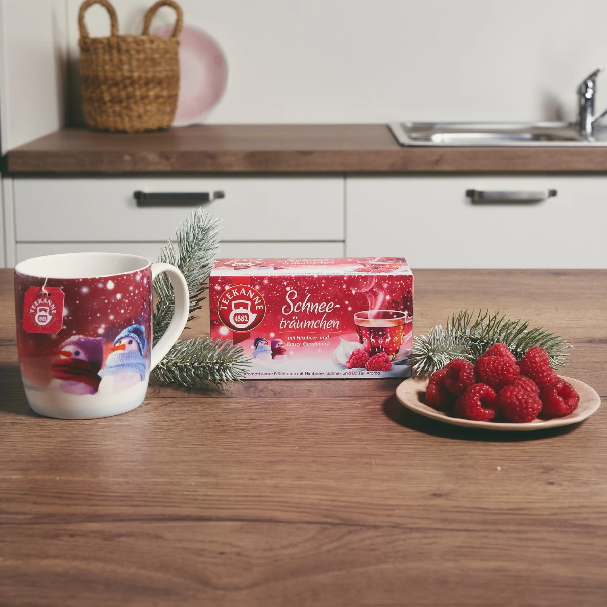 Das Schneeträumchen-Set mit Tasse steht mit einer Teeschachtel, einer festlichen Pinguintasse, einem Tannenzweig und Himbeeren vor einem modernen Küchenhintergrund auf einer hölzernen Küchentheke.