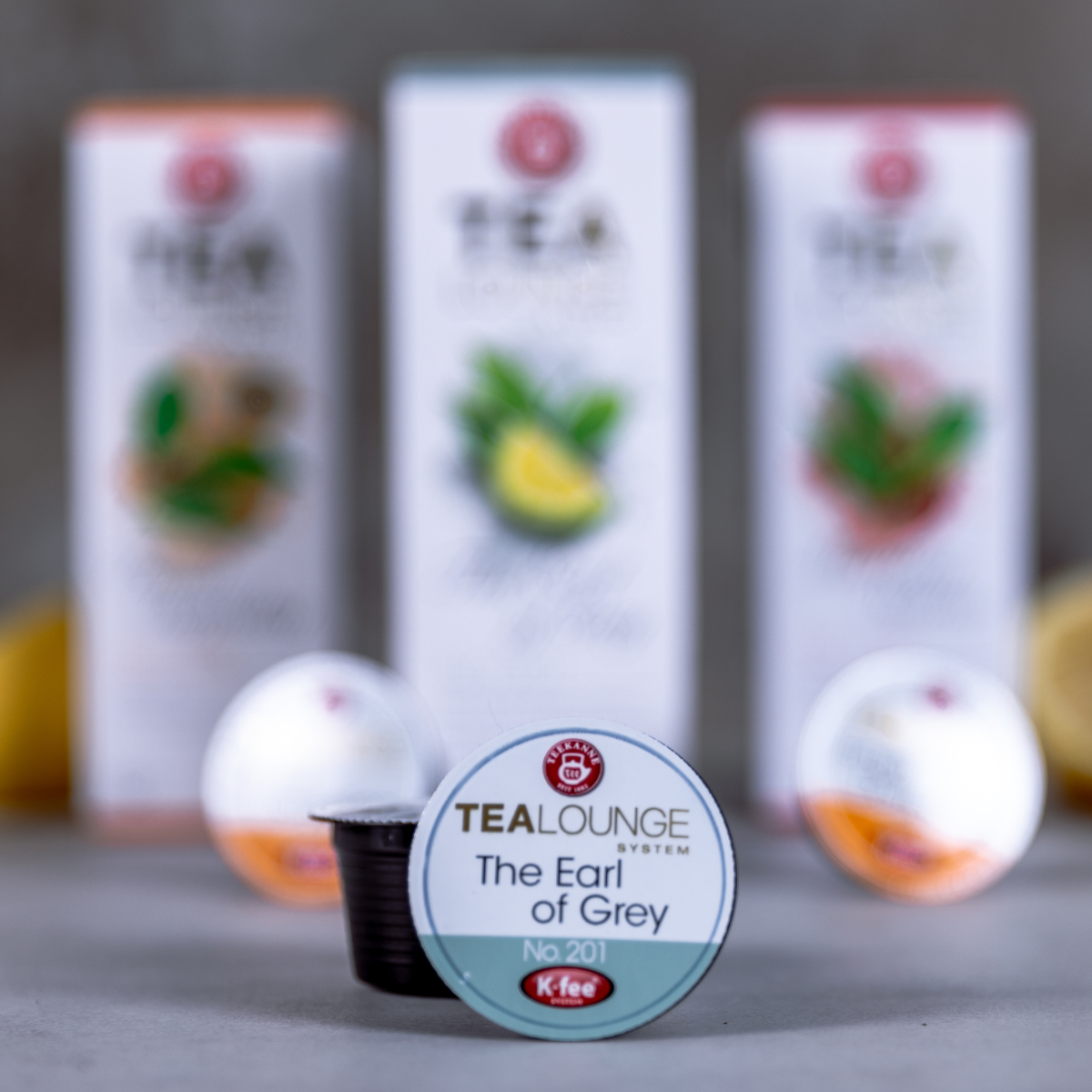 Im Fokus steht eine Nahaufnahme einer K-fee Teekapsel mit der Aufschrift „The Earl of Grey Nr. 201“, im Hintergrund sind verschwommene Teedosen und weitere Kapseln des TealoungeSystems zu sehen.