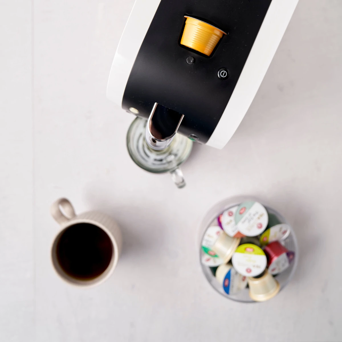 Draufsicht auf eine Kapselmaschine mit eingelegter goldener Kapsel, einem Glas unter dem Ausguss, einer beigen Tasse mit Moroccan Mint No. 681 Kaffee und einer Schale mit verschiedenen Kaffeekapseln auf einer hellen Oberfläche.