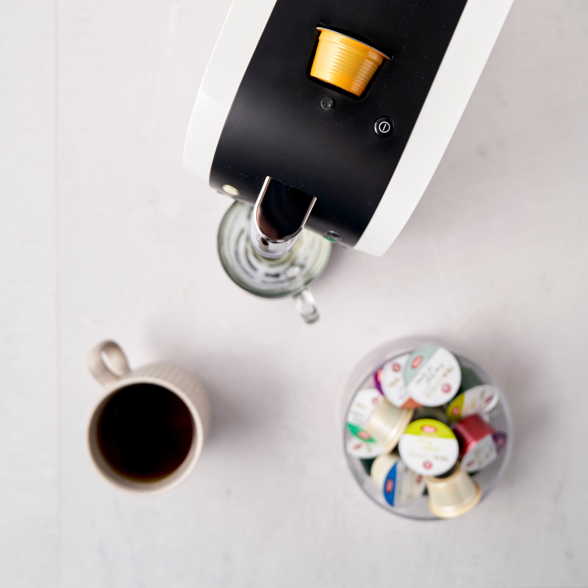 Eine Draufsicht auf eine Kapselmaschine Morning Sunrise Nr. 135 mit einer eingelegten goldenen Kaffeekapsel, einer Tasse Kaffee und einer Schale mit verschiedenen Kapseln auf einer hellen Oberfläche.