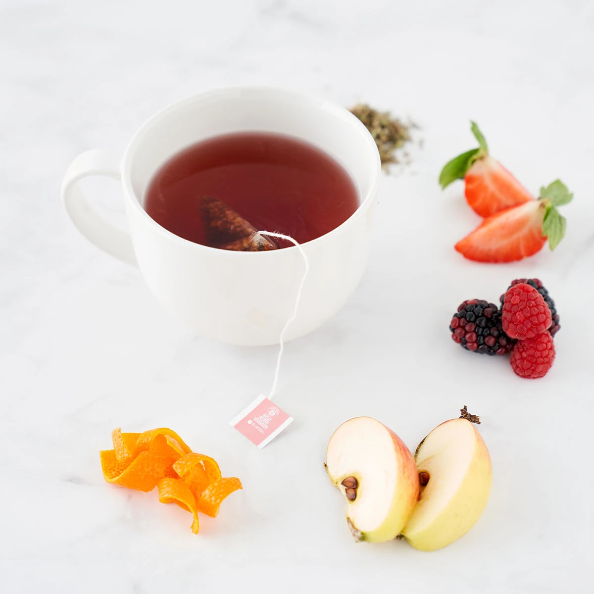 Eine Tasse Multivitamintee mit einem Teebeutel steht auf einer weißen Fläche, umgeben von geschnittenen Erdbeeren, Brombeeren, Himbeeren, Apfelscheiben, Orangenschalen und losen Teeblättern.