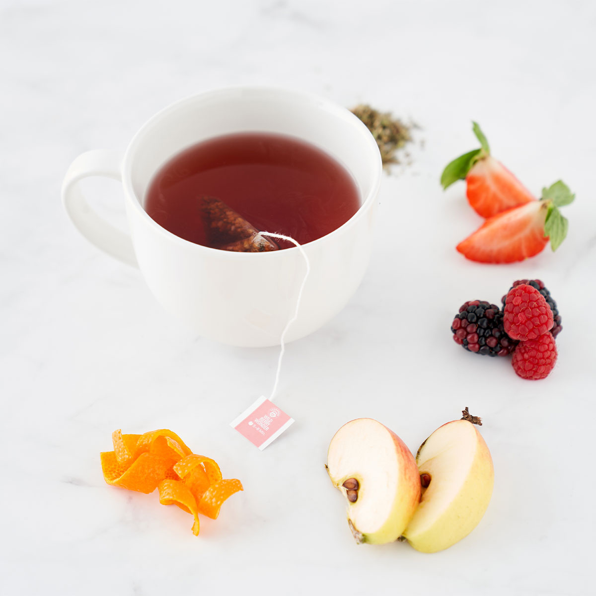 Eine weiße Tasse Früchte-Mischung-Tee mit Teebeutel steht auf Marmor, umgeben von frischen Erdbeeren, Brombeeren, Himbeeren, Apfelscheiben, Orangenschalen und losen TEEKANNE Früchte-Mischung-Blättern für puren Genuss.