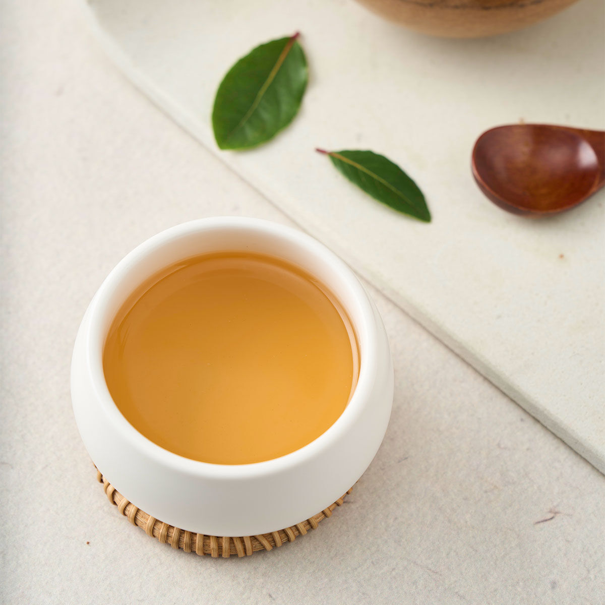 Eine weiße Keramiktasse, gefüllt mit Bio Grüner Tee, der seine hellgoldene Farbe zur Schau stellt, ruht auf einem gewebten Untersetzer neben zwei grünen Blättern und einem Holzlöffel auf einer hellen Oberfläche.