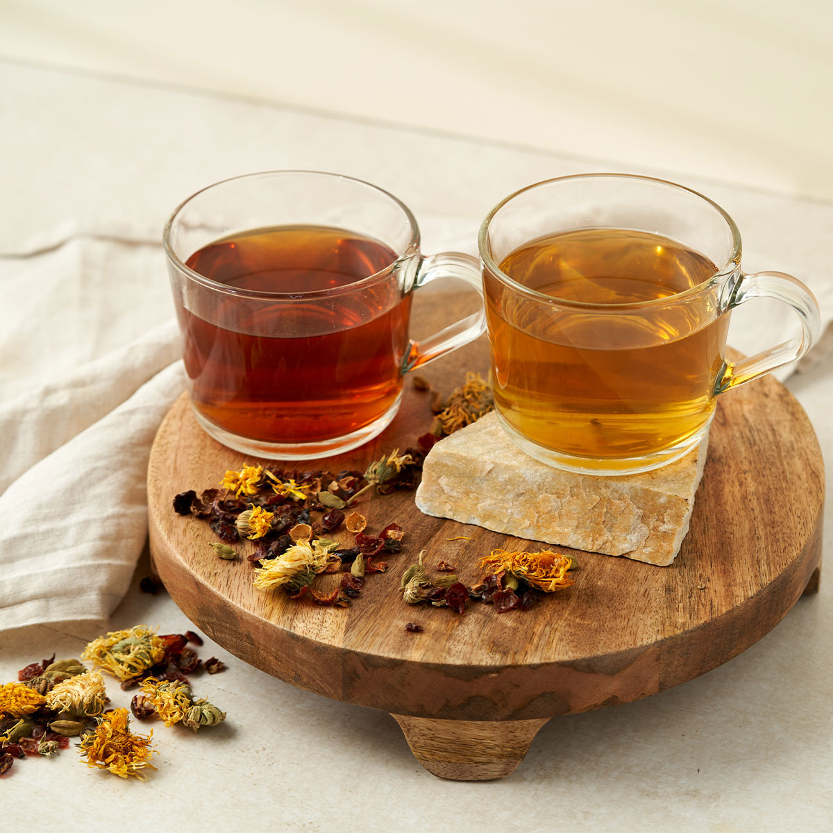 Zwei Glastassen mit BIO Basisch & Vital Tee, eine dunkelrote und eine hellgelbe, stehen auf einem runden Holzständer. Trockenblumen, Pfefferminze und Kräuter sind daneben verstreut, daneben liegt ein helles Tuch.