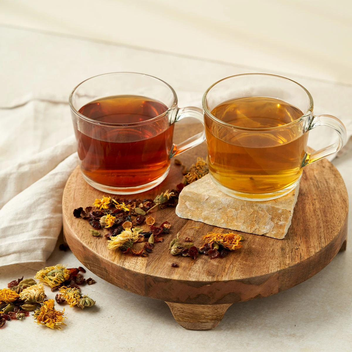 Zwei Glastassen mit BIO Basisch & Vital Tee, eine dunkelrote und eine hellgelbe, stehen auf einem runden Holzständer. Trockenblumen, Pfefferminze und Kräuter sind daneben verstreut, daneben liegt ein helles Tuch.