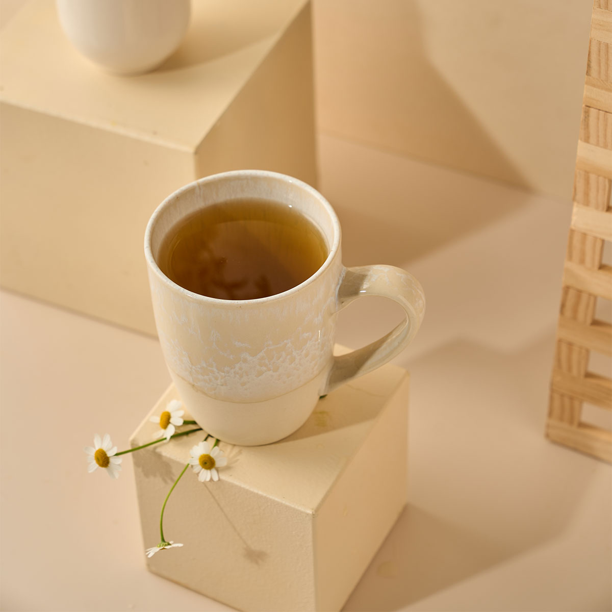 Eine beige Keramiktasse gefüllt mit Kamille-Kräutertee steht auf einem cremefarbenen Würfel neben weißen Gänseblümchen, mit neutral getönten Blöcken und sanfter Beleuchtung im Hintergrund.