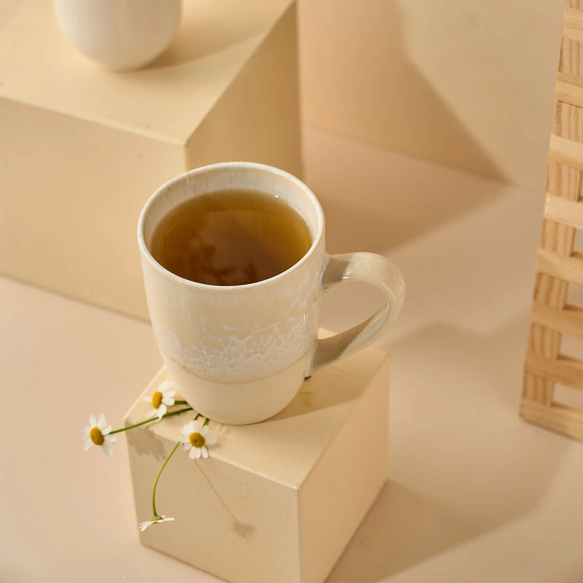 Eine cremefarbene Keramiktasse mit Kamille 100er Großpackung Tee steht auf einem beigen Würfel, umgeben von kleinen Gänseblümchen. Die Szene ist vor neutralen geometrischen Formen und sanfter Beleuchtung im Hintergrund gestaltet.