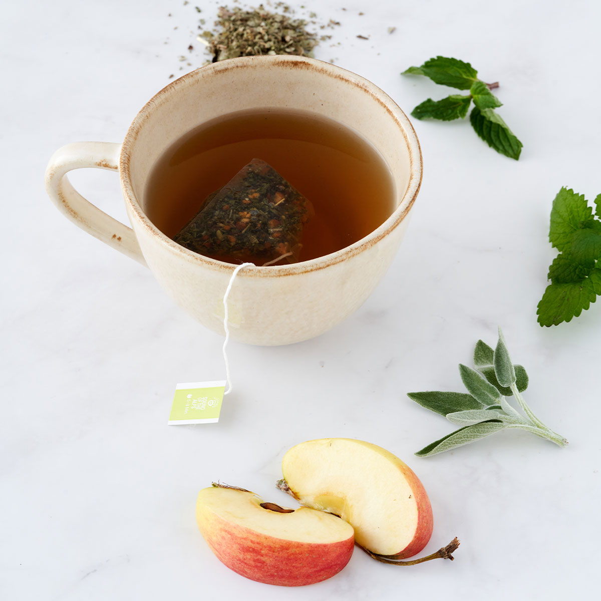 Eine Tasse Kräutertee mit einem Teebeutel darin ist umgeben von losen Blättern der Früchte-Mischung Familienpackung, frischer Minze, Salbei und roten Apfelscheiben auf einer weißen Marmoroberfläche.