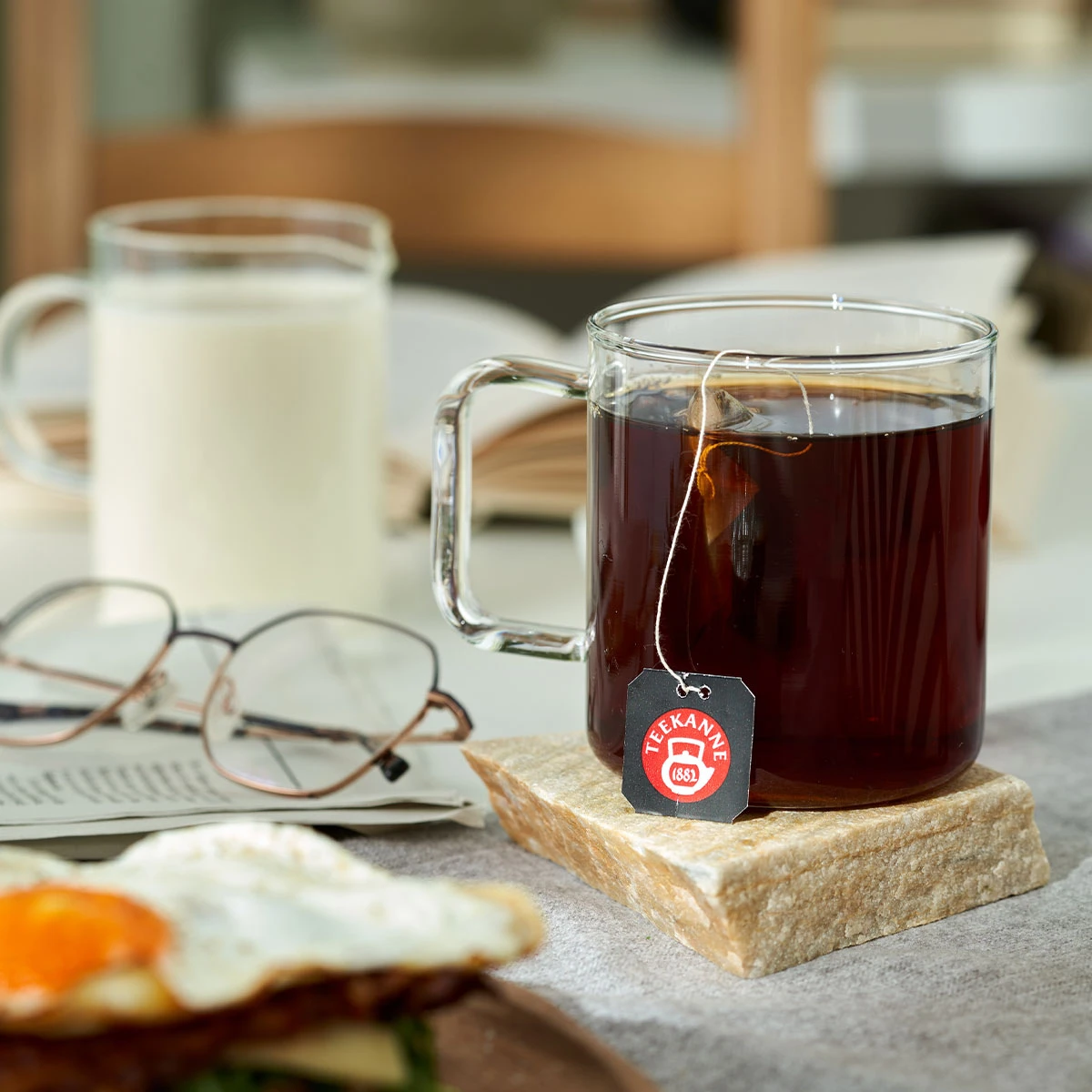 Eine Glastasse mit Ostfriesen Teefix Tasse 50er steht auf einem Steinuntersetzer neben einem Glas Milch, einer Brille und einem offenen Buch. Im Vordergrund ist ein Teller mit einem Spiegelei teilweise sichtbar.
