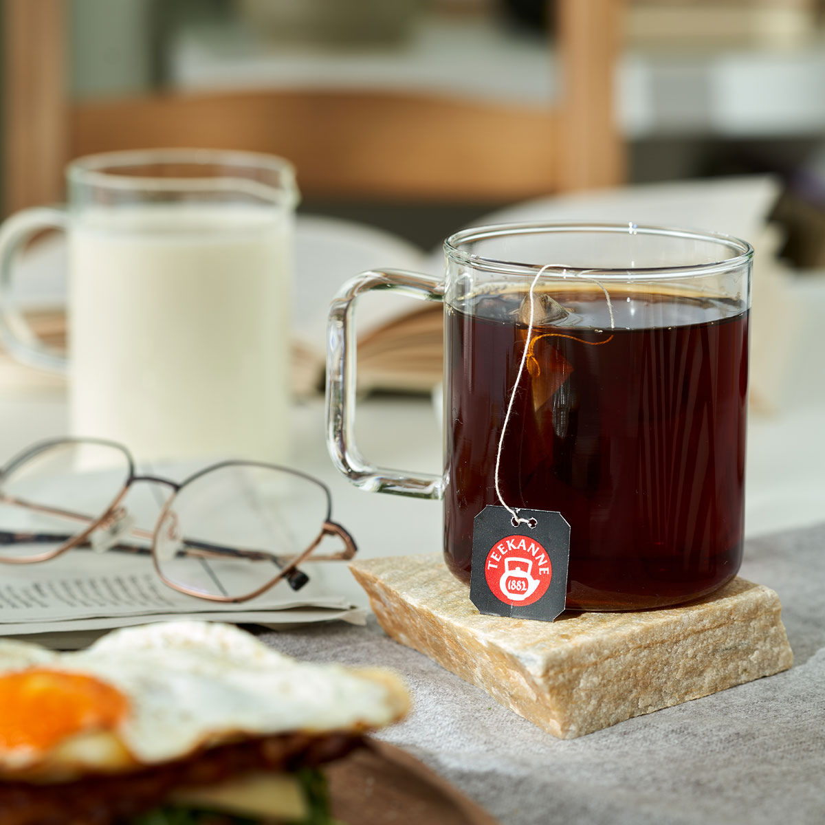 Ein Glas Bio Schwarzer Tee Earl Grey mit einem TEEKANNE-Teebeutel, ein Glas Milch, eine Brille, ein aufgeschlagenes Buch und ein Teller mit einem Spiegelei stehen einladend auf dem Tisch.