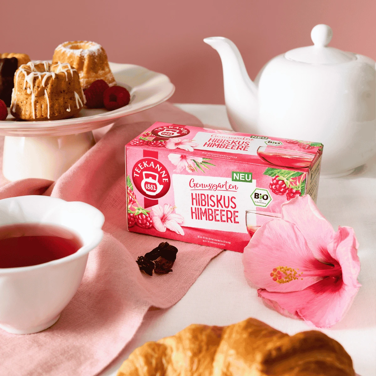 Auf einem Tisch steht eine Schachtel „Bio Hibiskus Himbeere“ neben einer Tasse Tee, einem Croissant, einer rosa Hibiskusblüte, einer Teekanne und kleinen Kuchen im Hintergrund.