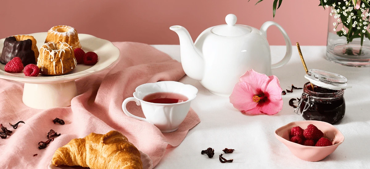 Auf einer rosa-weißen Tischdecke stehen eine weiße Teekanne, gefüllt mit Bio-Hibiskus-Himbeer-Tee, eine Tasse mit duftendem Tee, ein Croissant, Kuchen, Himbeeren, eine rosa Blume und ein Glas Marmelade; im Hintergrund steht eine Vase mit Blumen.
