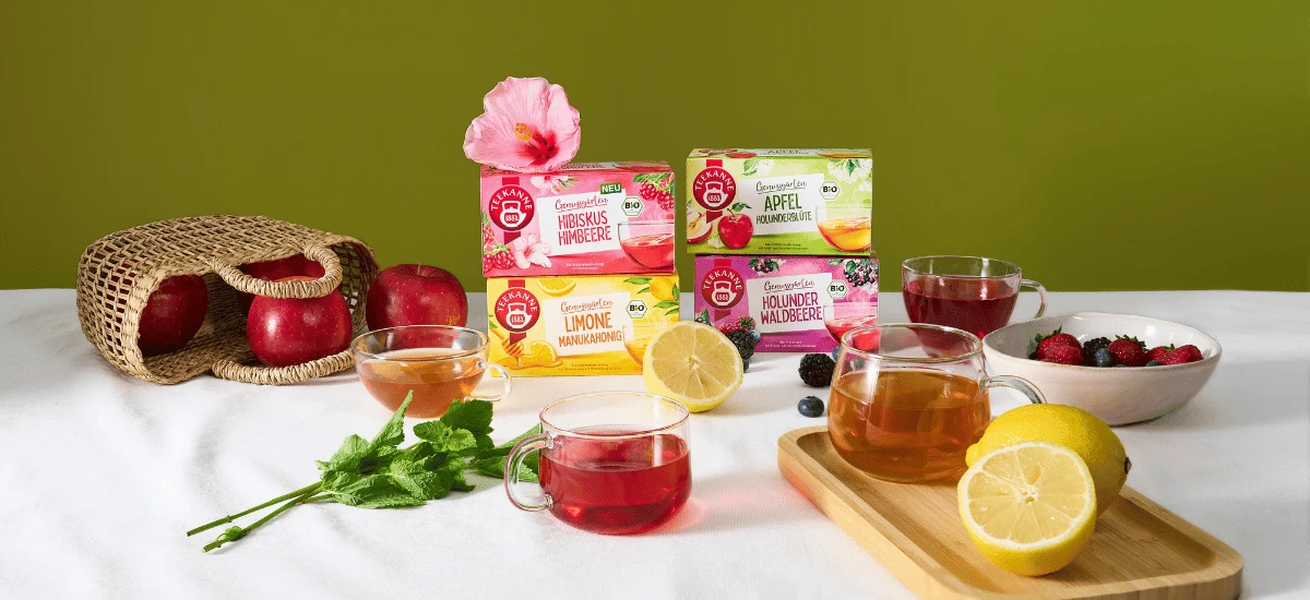 Drei durchsichtige Teetassen, gefüllt mit Kräutertee – unter anderem in den Geschmacksrichtungen Apfel und Bio-Hibiskus-Himbeere –, verschiedene Teedosen, frische Äpfel, Zitronenscheiben, Beeren wie Himbeeren und Minze sind auf einem weißen Tisch vor grün