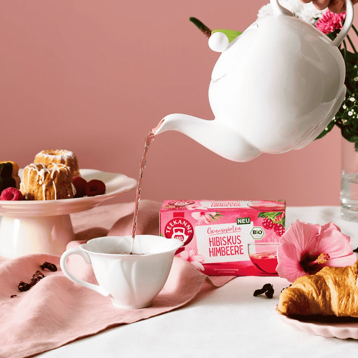 Eine weiße Teekanne gießt rosa Tee in eine Teetasse neben einem Croissant, verschiedenen Backwaren, einer Hibiskusblüte und einer Schachtel „Bio Hibiskus Himbeere“-Tee mit Noten von Himbeere und Apfel, die alle auf einem rosa-weißen Tisch angerichtet sind