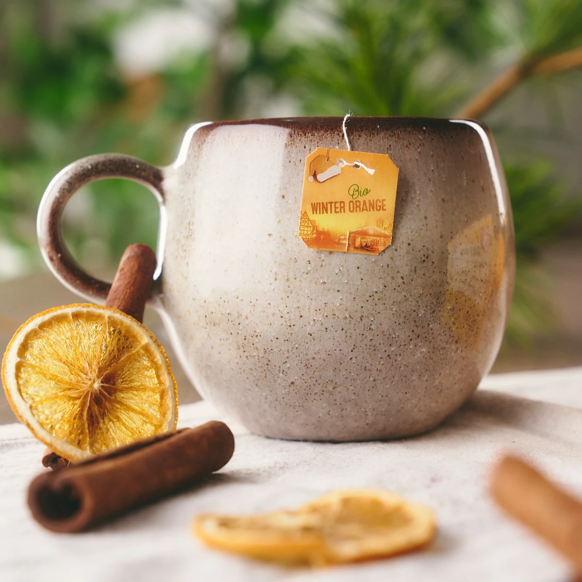 Eine Bio Winter Orange Keramiktasse mit Teebeutel steht auf einem Tisch, umgeben von Zimtstangen und getrockneten Orangenscheiben. Im Hintergrund sind verschwommene grüne Pflanzen zu sehen.