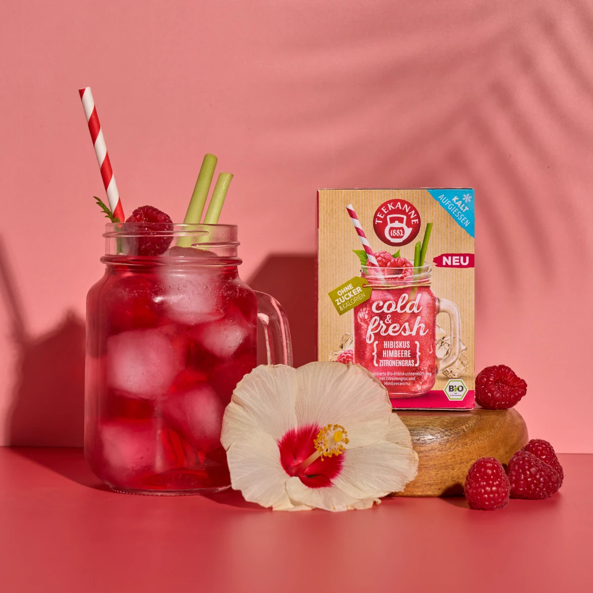 Ein Einmachglas mit rosa Eistee aus kaltem und frischem Hibiskus-Himbeere-Zitronengras, Himbeeren und Minze mit einem gestreiften Strohhalm steht neben einer Teebox, Himbeeren und einer blassen Hibiskusblüte auf einem rosa Hintergrund.