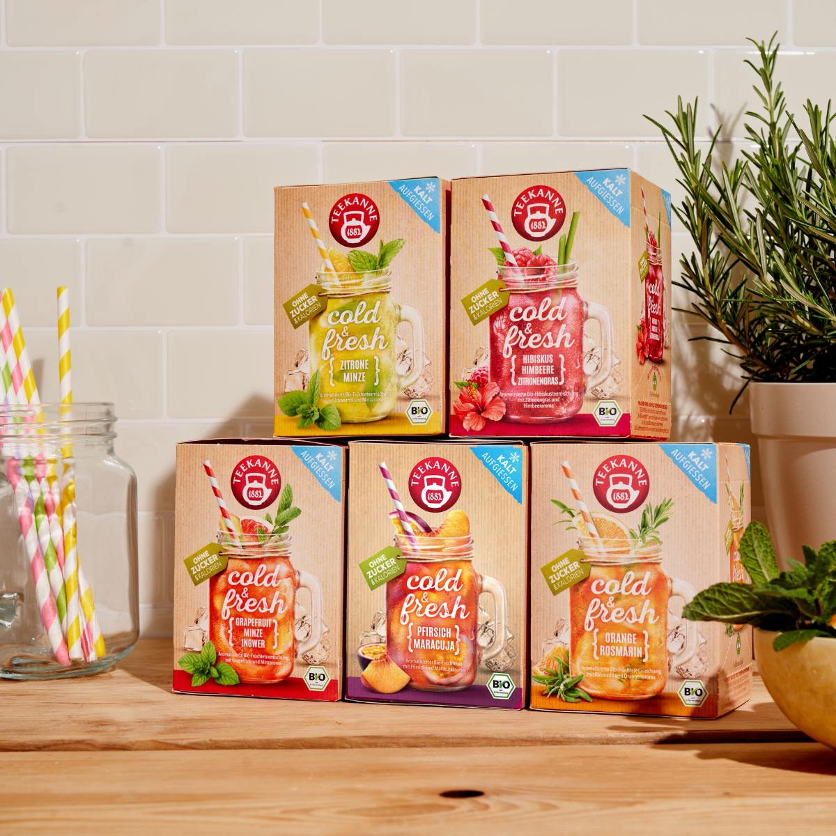Fünf Schachteln mit kaltem und frischem Mix & Match-Tee – Ingwer, Pfirsich, Zitrone – sind mit gestreiften Strohhalmen, Rosmarin und Minze auf einer Theke neben einer gefliesten Küchenwand gestapelt.