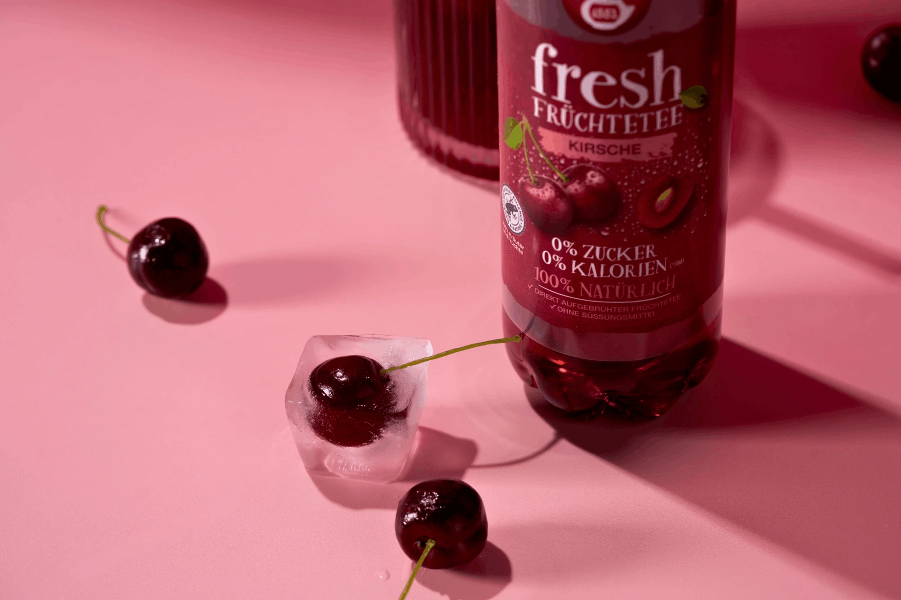 Ein Fresh Kirsche Sixpack steht auf einer rosa Fläche, umgeben von frischen Kirschen, einem Eiswürfel mit einer Kirsche darin und weiteren Flaschen aus der Packung, die teilweise sichtbar sind.