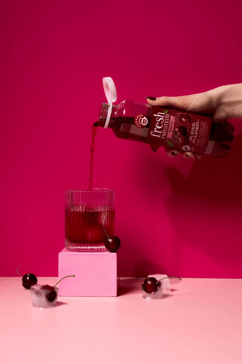 Eine Hand gießt Fresh Kirsche Sixpack roten Früchtetee aus einer Flasche in ein Glas auf einem rosa Block. Eiswürfel und Kirschen liegen daneben, alles vor einem kräftigen rosa Hintergrund.