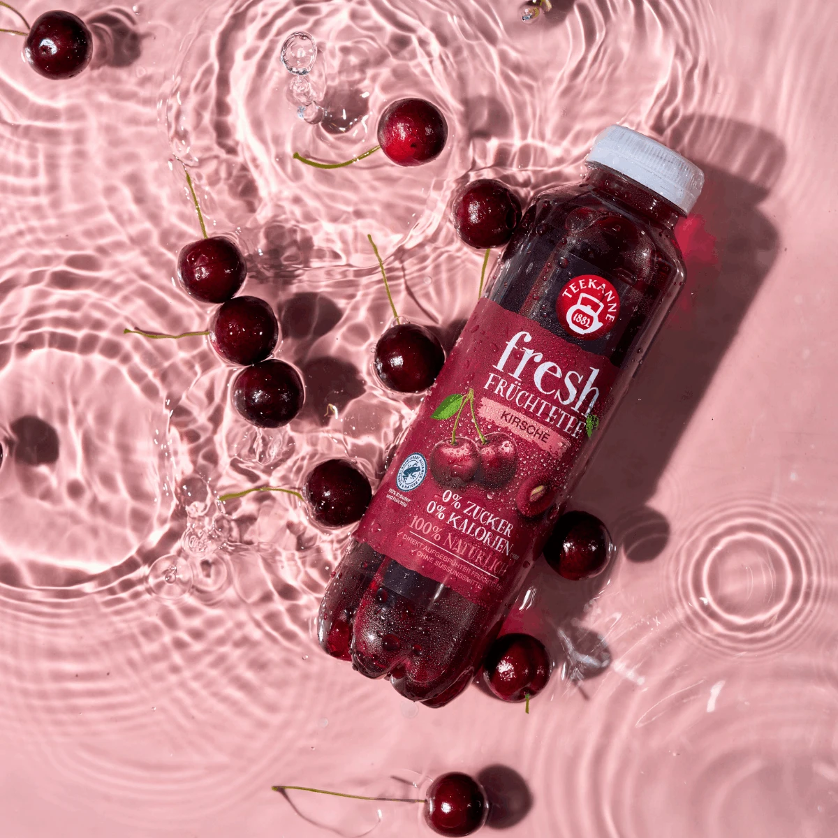 Eine Flasche Fresh Kirsche Sixpack Eistee schwimmt in rosafarbenem Wasser mit ganzen Kirschen und Plätschern. Auf dem Etikett stehen 0% Zucker, 0% Kalorien und 100% natürliche Zutaten - ideal für ein erfrischendes Sixpack.