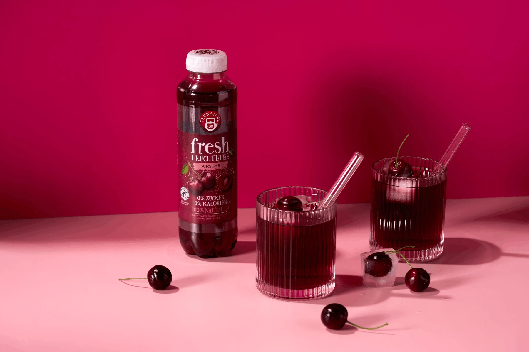 Eine Fresh Kirsche Sixpack-Flasche steht neben zwei Gläsern mit Eis, Kirsche und Saft auf einer rosafarbenen Fläche mit hellrosa Hintergrund. Lose Kirschen sind um die Gläser herum verstreut.