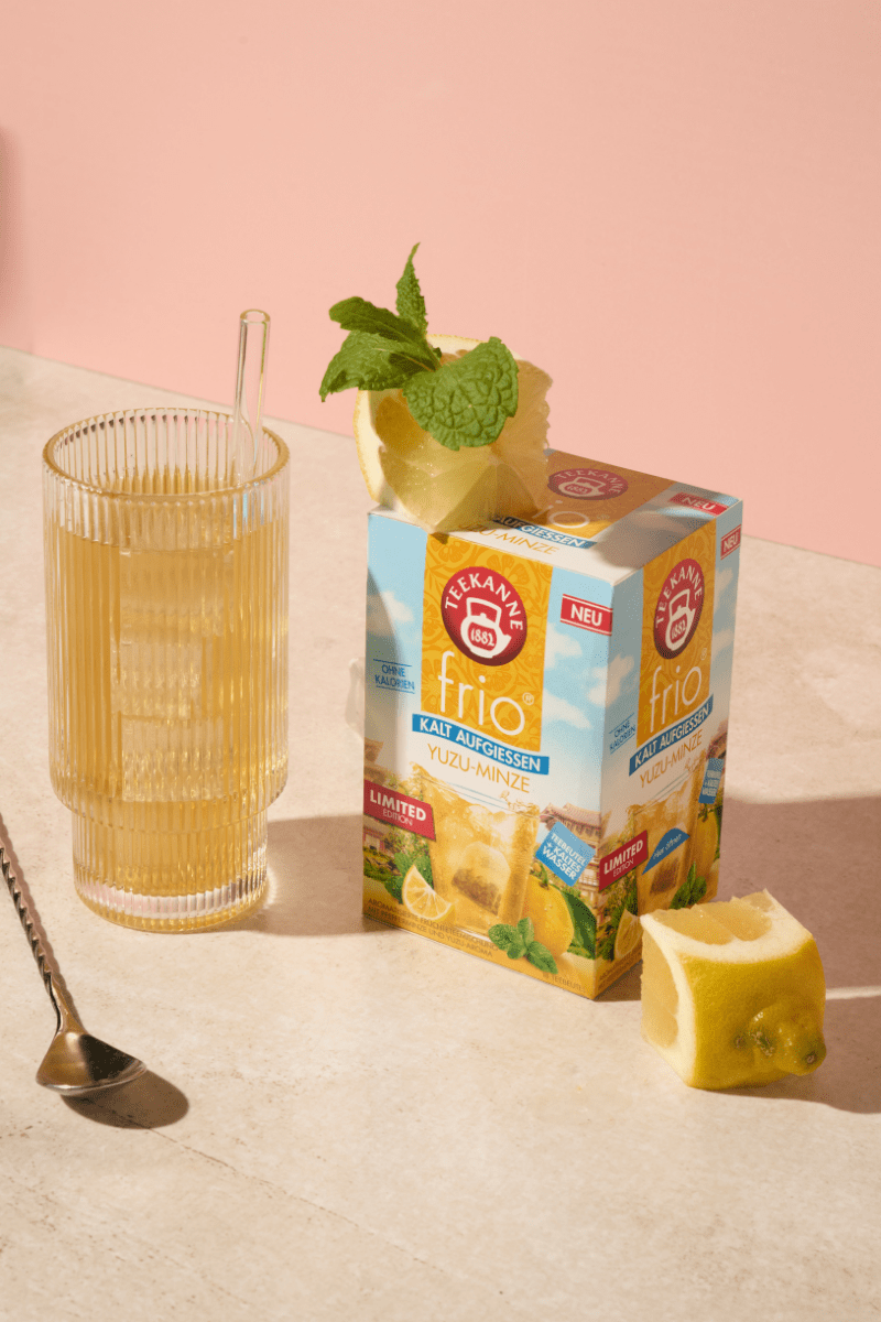 Eine Packung TEEKANNE frio Yuzu-Minze. Links daneben ein Glas mit aufgegossenem Tee, Eiswürfeln und Strohhalm. Dekoriert mit frisch angeschnittener Yuzu-Frucht.