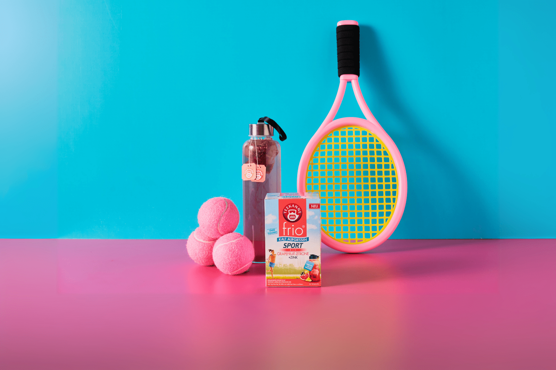 Eine Packung frio Sport Fit Grapefruit-Zitrone auf pinkem Untergrund und blauem Hintergrund. Daneben eine Glas-Sportflasche mit aufgegossenem Tee. Dazu ein Tennisschläger und Tennisbälle.