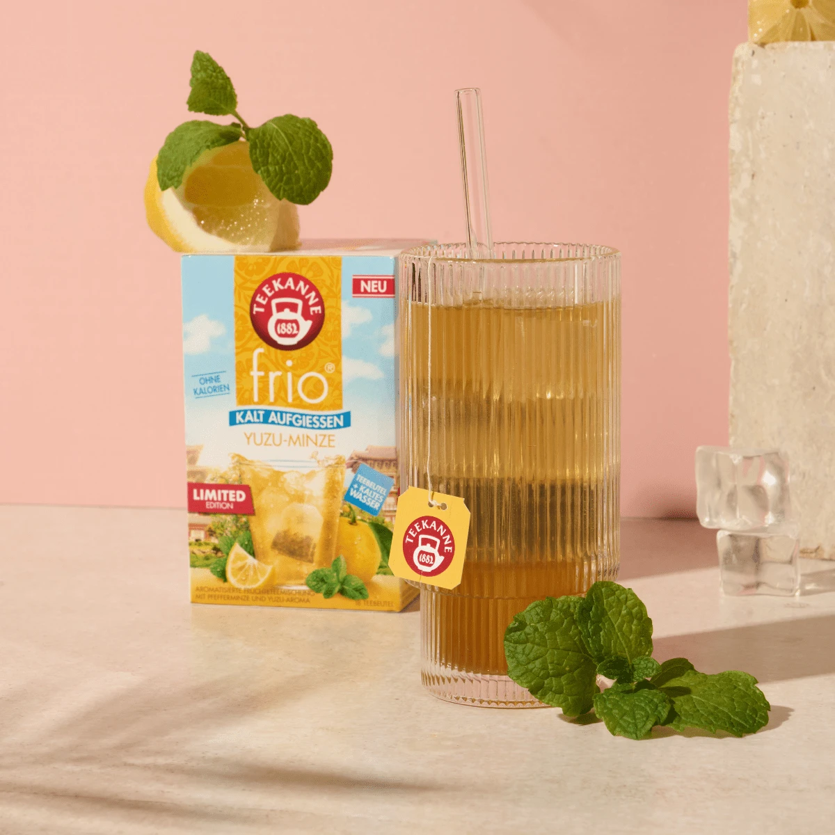 Eine Packung "frio" in der Geschmacksrichtung "Yuzu-Minze". Ein Tee zum kalt Aufgießen. Aufgegossen in einem Glas mit Strohhalm. Dekoriert mit frischer Yuzu-Frucht, Minze und Eiswürfeln.