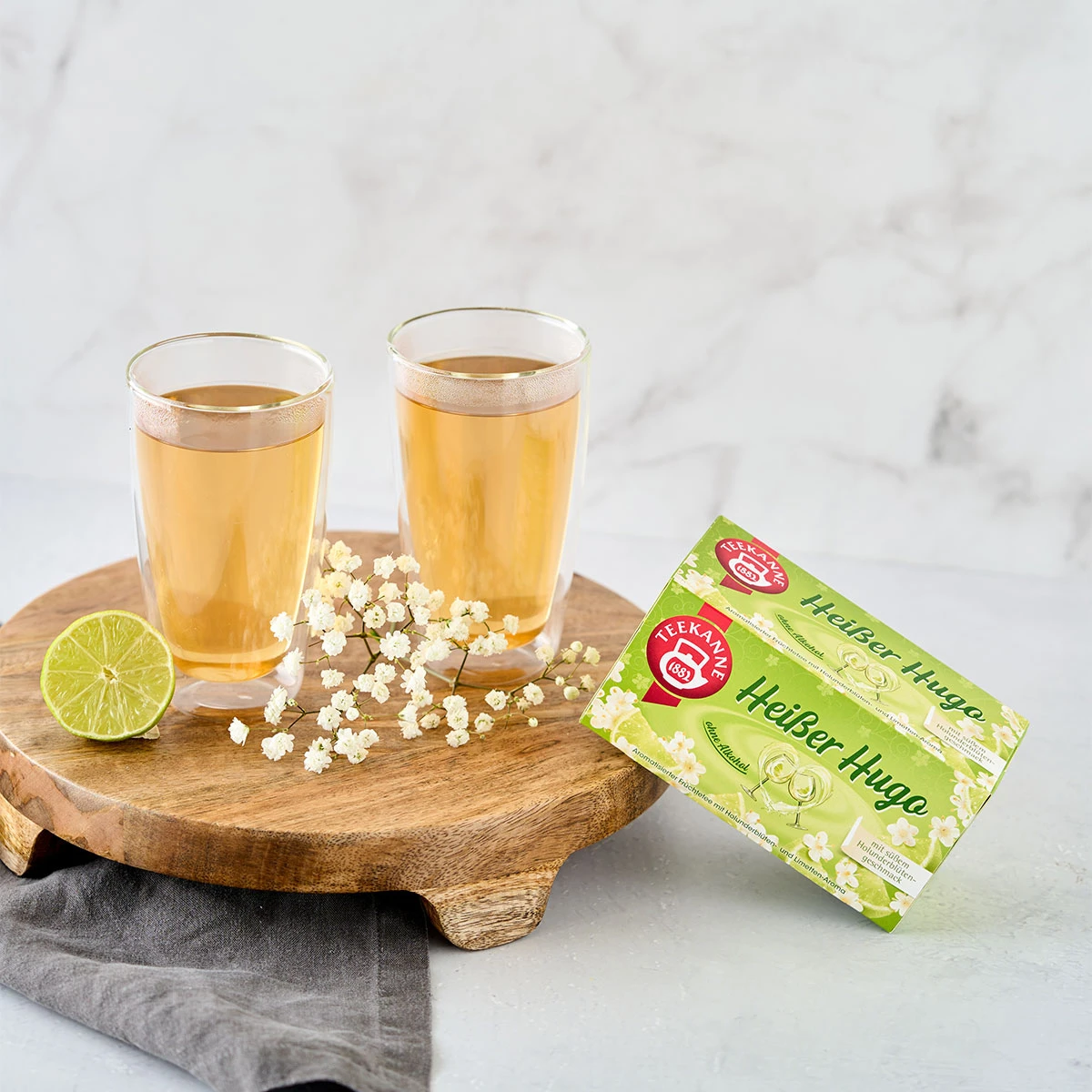 Zwei Gläser hellen Verführerische Früchtetees Mix & Match Tee stehen auf einem runden Holzbrett mit Limettenscheiben und kleinen weißen Blüten. Die Teebox steht daneben auf einer hellen Ablage vor einem Marmorhintergrund.