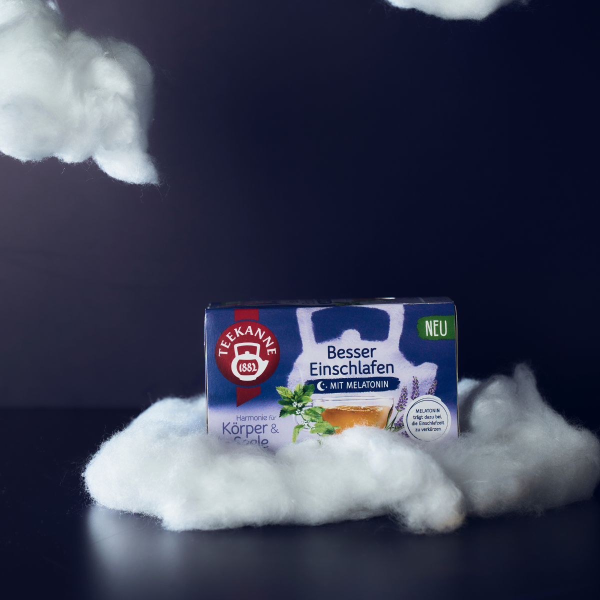 Eine Schachtel Besser Einschlafen Kräutertee mit Melatonin liegt zwischen weichen, wolkenartigen Requisiten vor einem dunklen Hintergrund, sorgt für Gleichgewicht für Körper und Seele und lädt zur Entspannung für erholsamen Schlaf ein.