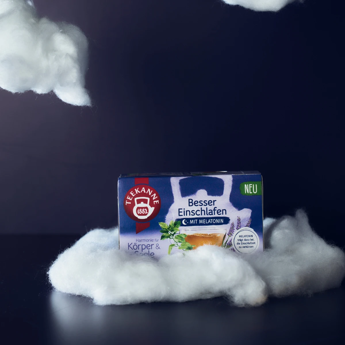 Eine Schachtel Besser Einschlafen Kräutertee mit Melatonin liegt zwischen weichen, wolkenartigen Requisiten vor einem dunklen Hintergrund, sorgt für Gleichgewicht für Körper und Seele und lädt zur Entspannung für erholsamen Schlaf ein.